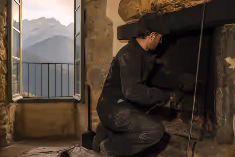 Ramoneur en tenue professionnelle inspectant un conduit de cheminée dans une maison en pierre du pays de Foix, avec vue sur les contreforts pyrénéens en arrière-plan